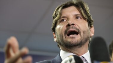 André do Prado, de cabelo castanho na altura do pescoço e barba, fala diante de microfones com a mão erguida. Ele veste terno escuro, camisa branca e gravata.