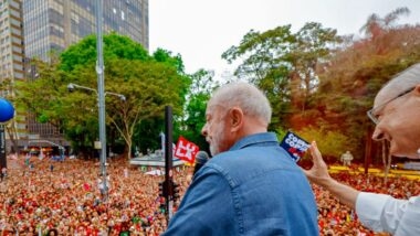 Lula, de camisa jeans, fala ao microfone de costas para a câmera, diante de uma multidão vestida de vermelho em na avenida Paulista. À direita, Geraldo Alckmin, de óculos e camisa branca, acena. Ao fundo, uma bandeira com o nome Lula.