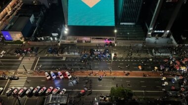 Vista aérea noturna da Avenida Paulista, em São Paulo, com dezenas de viaturas da Polícia Militar e uma van da Guarda Civil Metropolitana enfileiradas no asfalto, policiais a pé entre as faixas e pessoas com guarda-chuvas na calçada, na noite do segundo turno da eleição presidencial.